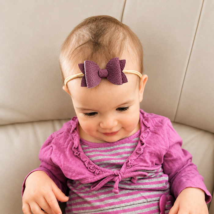 Baby wearing a purple headband with a bow, sitting on a beige couch.