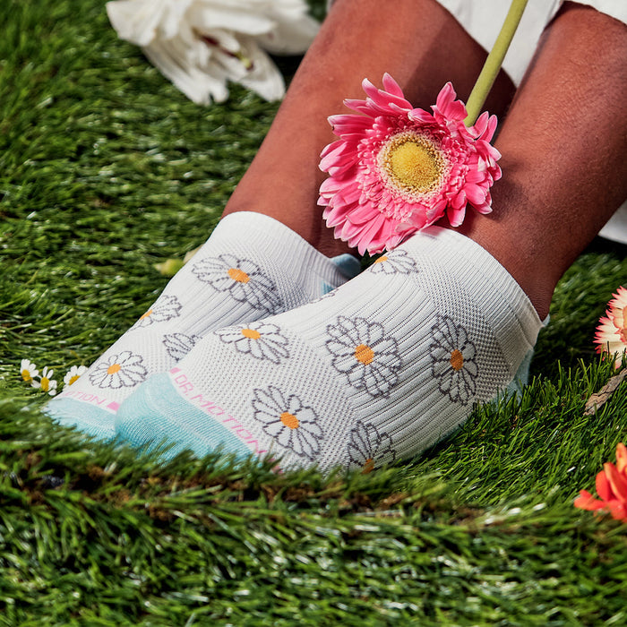 Ankle compression socks with a daisy pattern, worn on feet resting on grass, with a flower held between the toes.