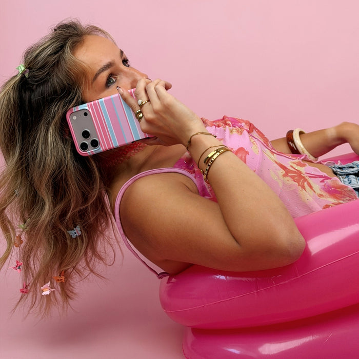 A woman reclining in a pink inflatable chair, against a solid pink background, holding a iphone with a pink, blue, and white striped phone case.