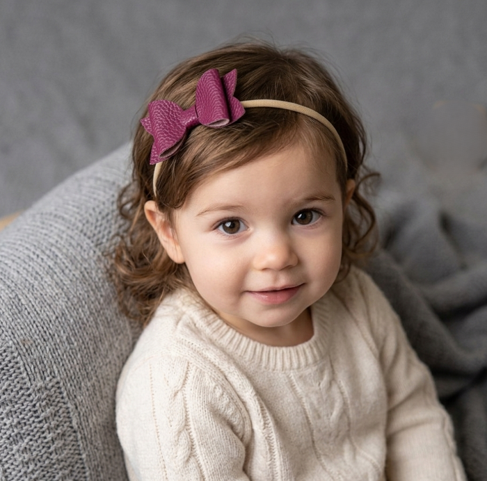 Toddler girl wearing a plum coloured bow headband, sitting on a grey surface.