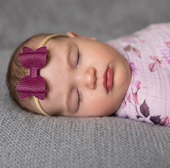 Newborn baby sleeping, wearing a purple bow headband and pink floral swaddle, laying on a grey blanket.