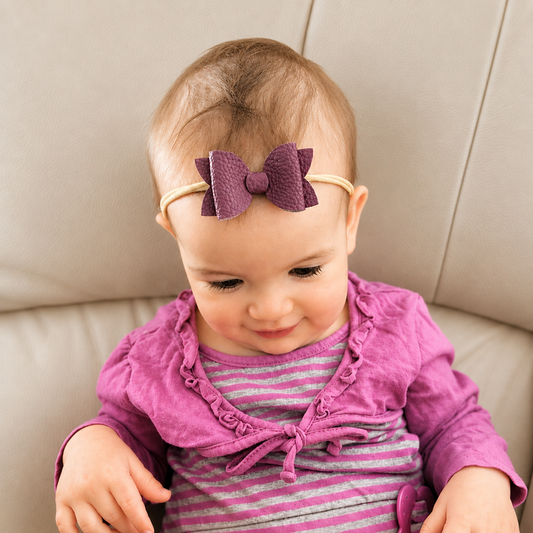Baby wearing a purple headband with a bow, sitting on a beige couch.