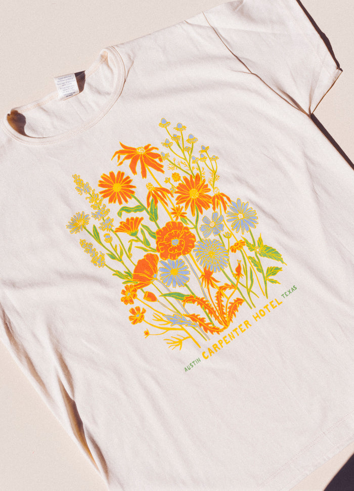 Photograph of beige cotton T-shirt with colorful floral print with text below it that reads 'CARPENTER HOTEL AUSTIN TEXAS' laying on top of beige background lit by natural sunlight