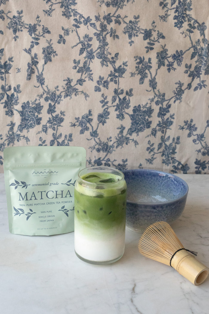 Glass of matcha with a bamboo whisk and a blue bowl, placed in front of the maman matcha powder pouch.