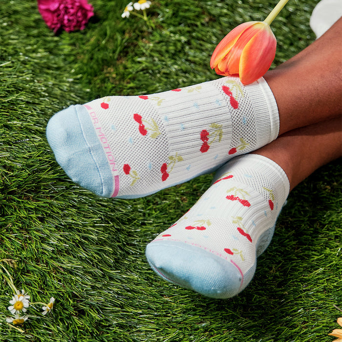 Ankle socks with a cherry pattern and light blue accents, resting on grass with a tulip nearby.