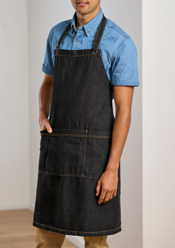 A man wearing a dark charcoal gray denim bib apron over a light blue short-sleeve button-down shirt, standing against a neutral off-white wall.