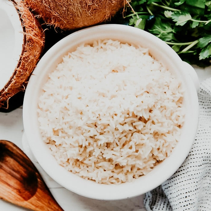 A bowl of cooked white rice with a sprinkle of cilantro on top, alongside a partially peeled coconut and a wooden spoon.