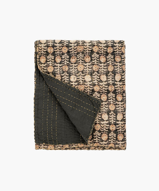 Folded throw blanket or quit with intricate patterns of beige circular floral motifs on a dark background. The corner reveals a contrasting black underside with yellow stitching. | Wildwood Home Co.