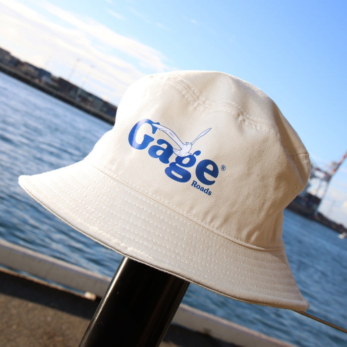 Cream bucket hat with 'Gage' logo on a metal pole by a waterfront