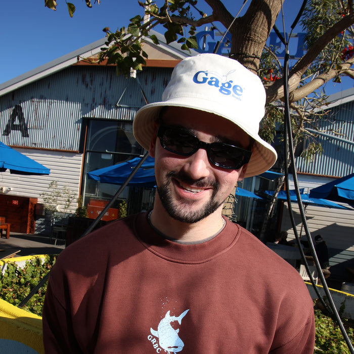 Man wearing a cream 'Gage' bucket hat and sunglasses, standing in front of a building with blue awnings.