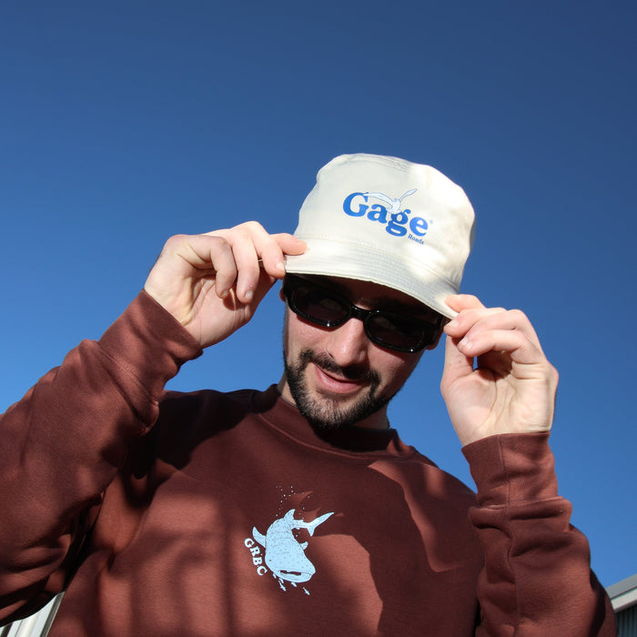 Man wearing a brown sweatshirt with a logo and a cream bucket hat with 'Gage' text against a blue sky.