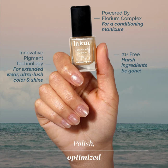A hand holds a bottle of LONDONTOWN Isla Bonita nail polish, showcasing a natural pink manicure with shimmering gold accents by the ocean. Text highlights Isla Bonita’s radiant color and nourishing formula.