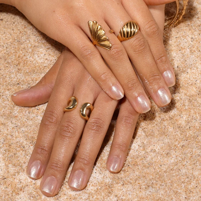 Two hands painted with LONDONTOWN’s Isla Bonita, an iridescent light pink nail polish, adorned with gold rings, rest gently on the sand.