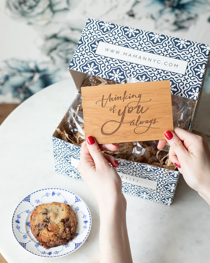 Person taking a wooden postcard engraved with “Thinking of You Always” in a calligraphic script from a blue-and-white gift box with cookies.
