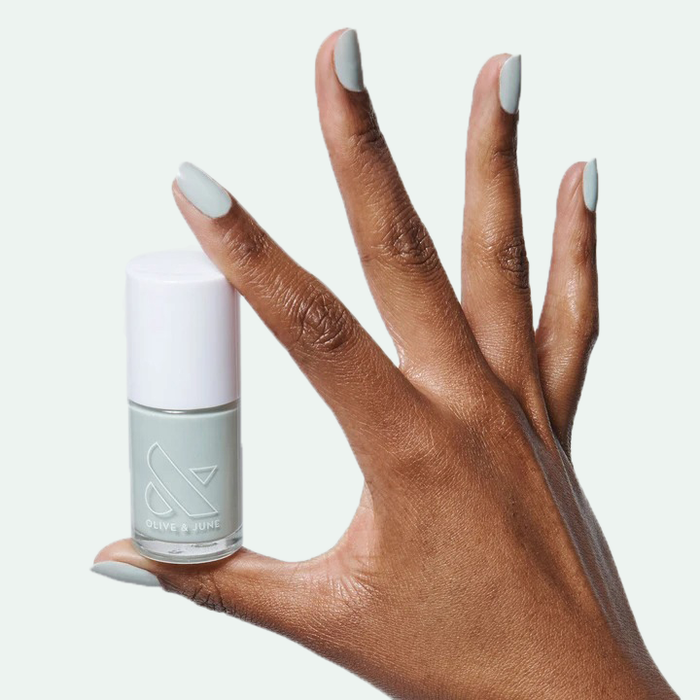 Hand holding a bottle of KMC sage green polish with matching manicure on medium brown skin tone for at-home mani