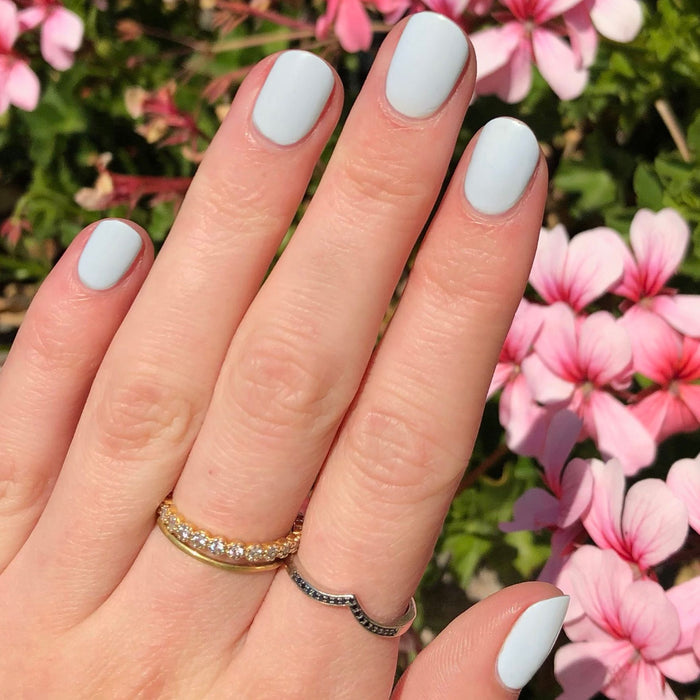 Hand with nails painted in soft pale blue KMC gel polish showing a smooth, elegant manicure outdoors near pink flowers