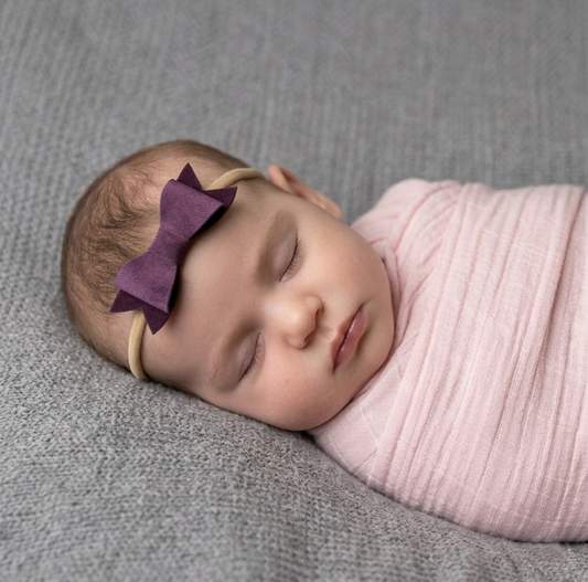 Newborn baby sleeping, wearing a purple bow headband, laying on a grey blanket.