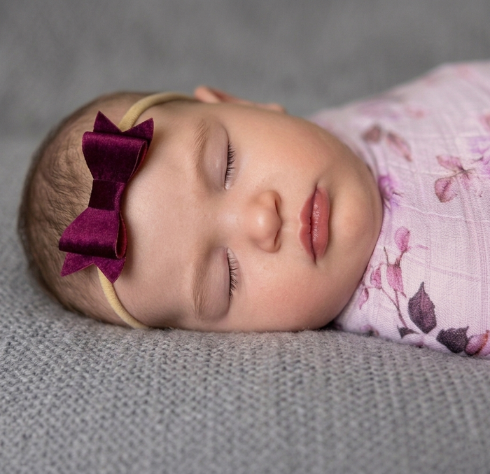 Newborn baby wrapped in floral swaddle, wearing a plum coloured bow headband and laying on a grey blanket.