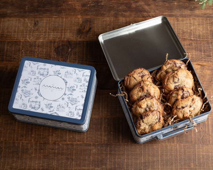 A open blue and white toile tin lunch box filled with chocolate chip cookies, with a closed lunch box next to it.
