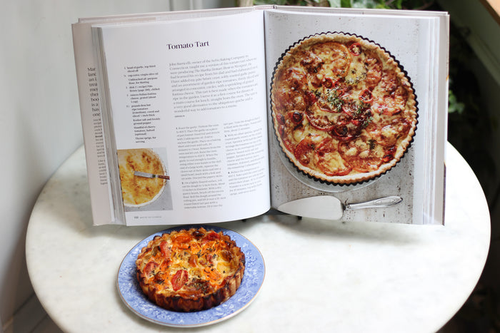 Opened Martha Stewart cookbook titled "THE COOKBOOK" showing a tomato tart recipe with a real tart on a plate below.