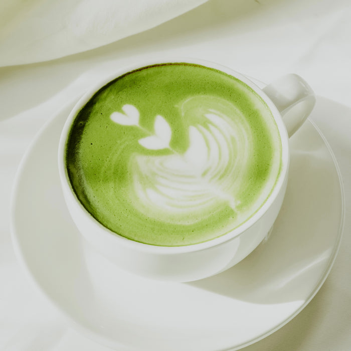 A cup of matcha latte with green foam art in the shape of a heart on top, served in a white cup and saucer.