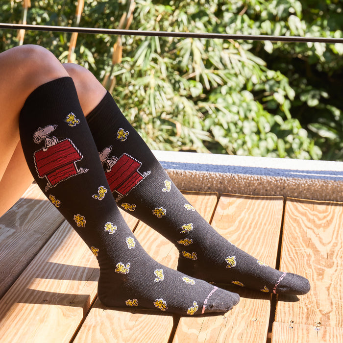 Knee-high black compression socks featuring Snoopy on a doghouse and yellow flowers.
