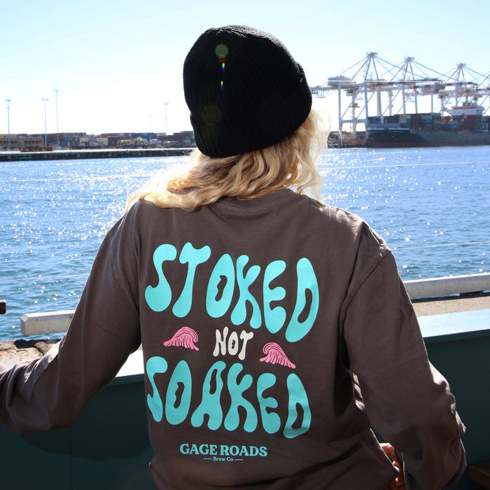 Person wearing a grey sweatshirt with 'Stoked Not Soaked' text by a waterfront.