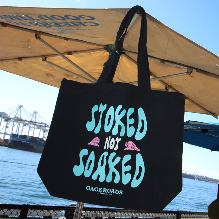 Black tote bag with 'Stoked Not Soaked' text under a large umbrella by a waterfront.