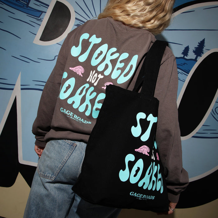 Person wearing a hoodie and carrying a tote bag with 'Stoked Not Soaked' text, standing in front of a colorful mural.