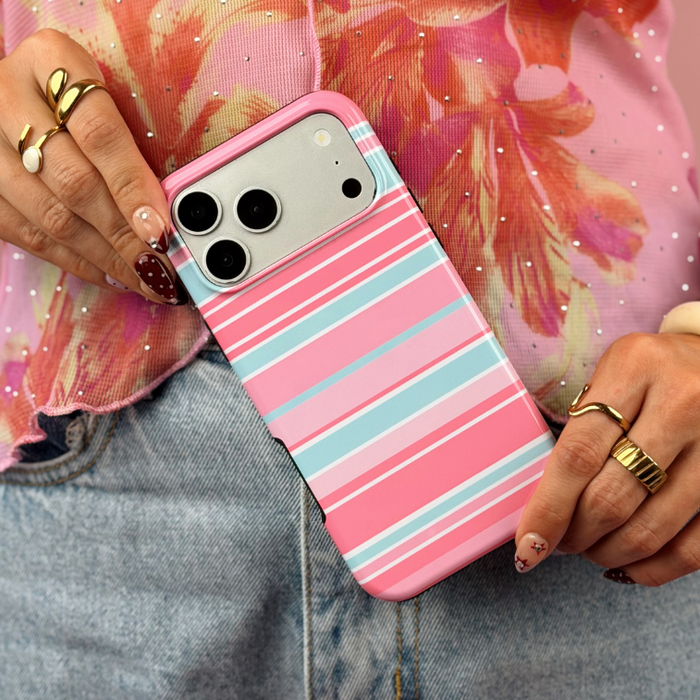 A close-up of a person holding a iphone in a pink, blue and white striped case, wearing a pink floral top and light blue jeans against a solid pink background.