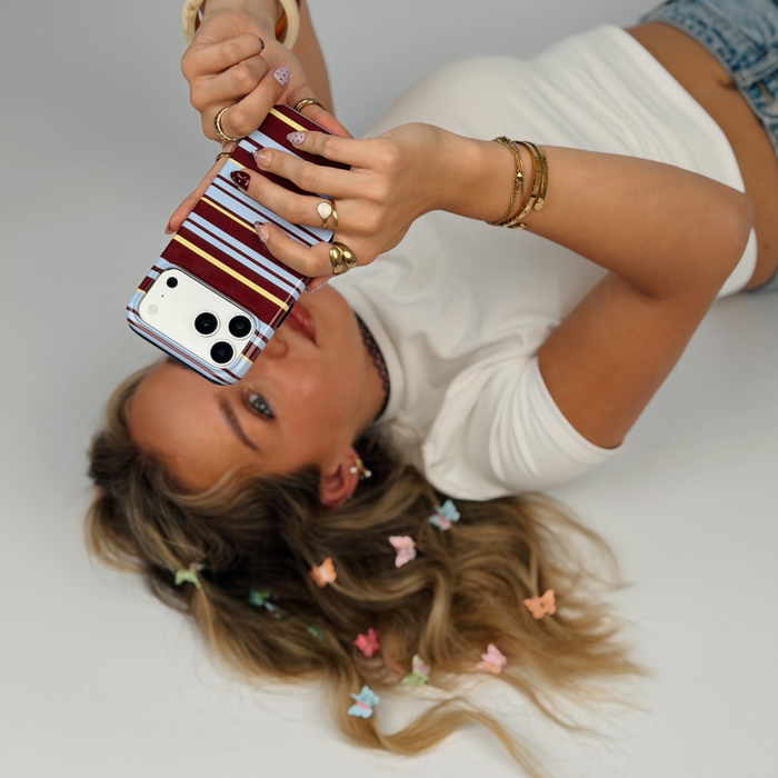 A woman lying on a white floor holds an iphone with a brown, blue and yellow striped phone case. She is wearing a white top, denim shorts, and multiple gold rings.
