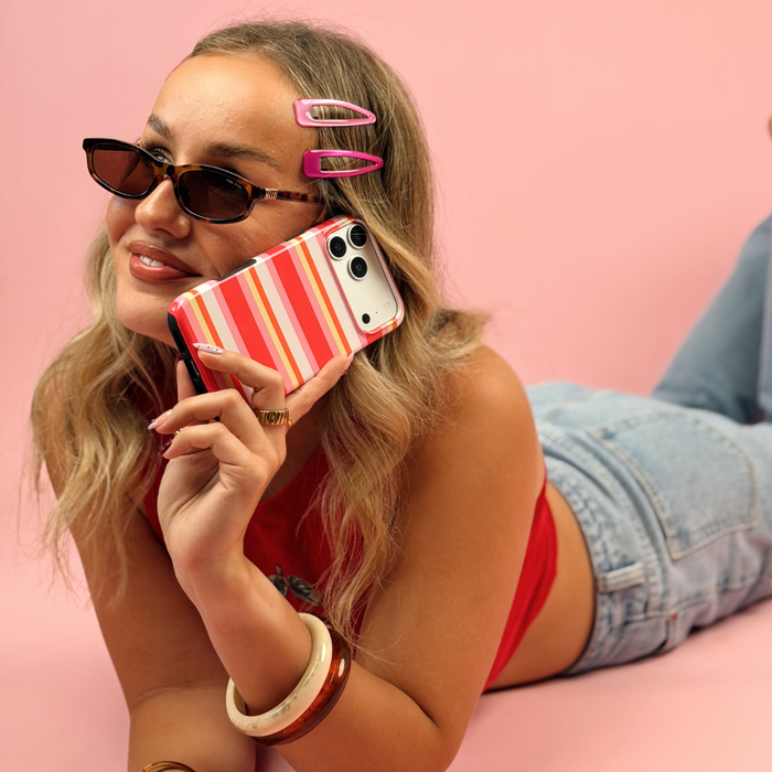 A woman lying on a pink surface wearing cat-eye sunglasses, pink hair clips, a red top, and denim shorts while holding a striped hot pink, orange, yellow, and white.