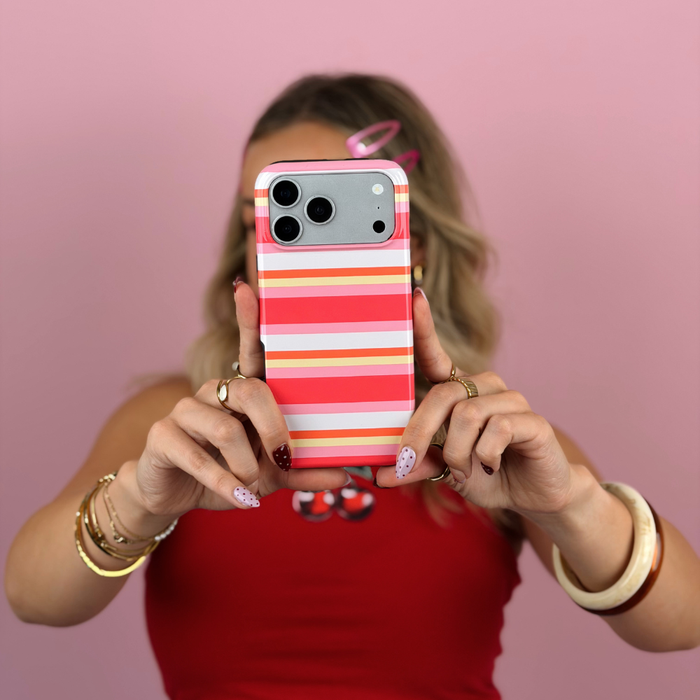 A woman holding a smartphone with a   pink, orange, yellow, and white phone case in front of her face, wearing a red top and gold jewellery against a pink background.