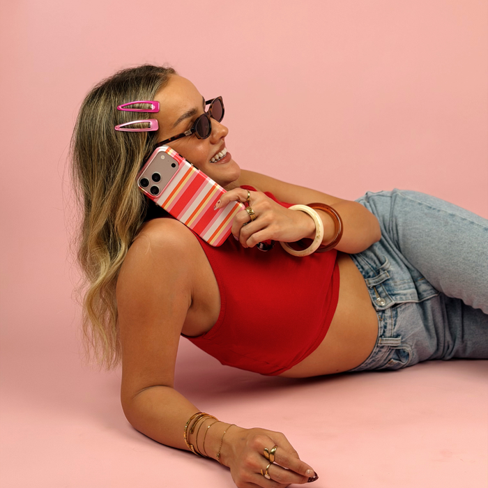 A woman lying on a pink studio floor wearing a red crop top, light-wash denim jeans, sunglasses, and pink hair clips, holding a phone with a striped pink, orange, yellow, and white case.