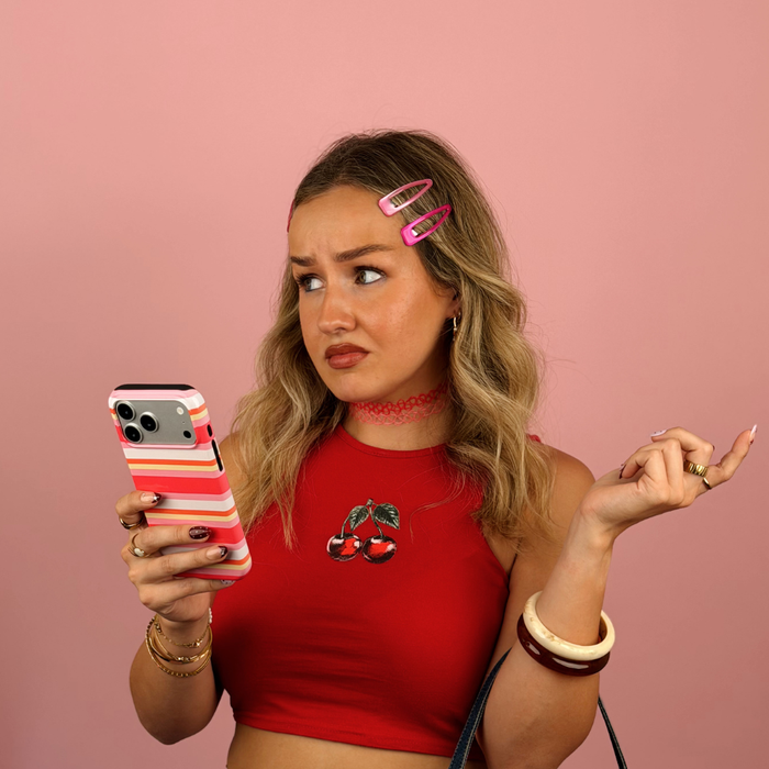 A woman with blonde hair in a red crop top with a cherry graphic, holding a striped  pink, orange, yellow, and white phone case. She is posing against a solid pink background with pink hair clips.
