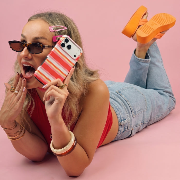 A woman lying on her stomach against a solid pink background, holding an iPhone with a pink, orange, yellow, and white case. She is wearing sunglasses, a red top, light-wash blue jeans, and bright orange platform shoes.