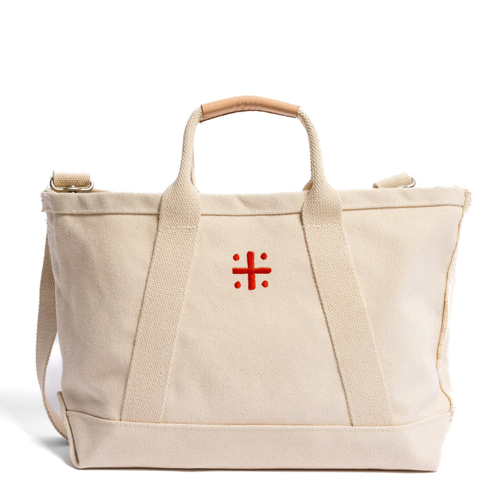 A beige canvas tote bag with leather handles and a red cross logo on the front.