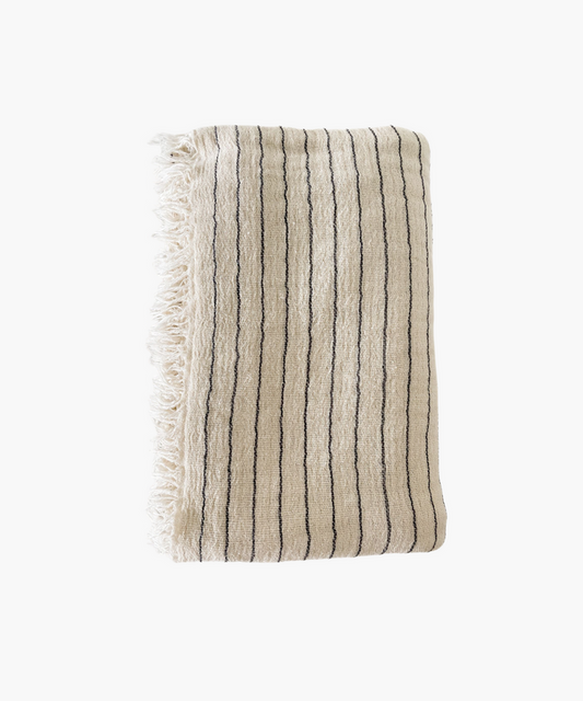 A folded beige throw blanket with thin black stripes and fringed edges. The fabric appears textured and soft, evoking a sense of warmth and comfort. | SW Home