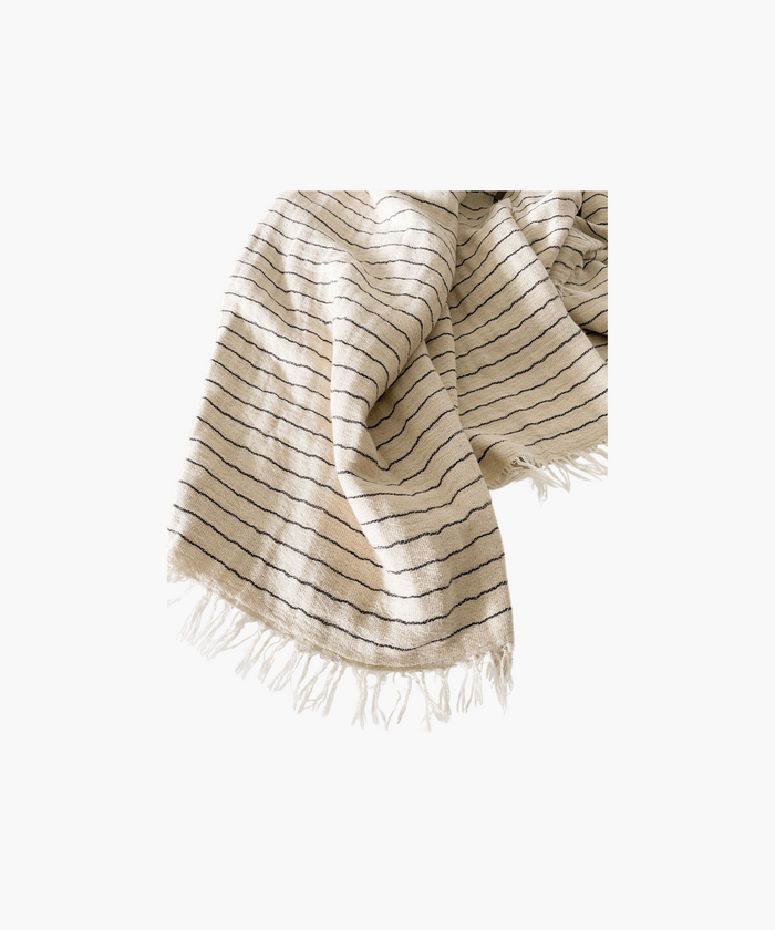 A folded beige throw blanket with thin black stripes and fringed edges. The fabric appears textured and soft, evoking a sense of warmth and comfort. | SW Home